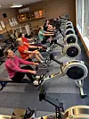 Menschen rudern nebeneinander auf Indoor-Rudergeräten in einem Trainingsraum.