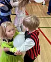 Zwei verkleidete Kinder tanzen eng umklammert mit einem weißen Luftballon in einer Turnhalle.