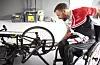 Mann im Rollstuhl beugt sich zu einem Handbike in einer Werkstattumgebung.