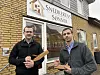 Zwei Männer halten Holzmuster vor dem Showroom von Snedkerens Sønner in einer Backsteinfassade.