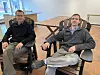 Zwei Männer sitzen in massiven Holzsesseln in einem hellen Ausstellungsraum.