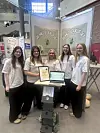 Fünf Schülerinnen stehen an einem Messestand mit Plakaten, Urkunde und Laptop.