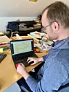 Person sitzt an überladenem Schreibtisch und arbeitet an Lenovo-Laptop in Dachbüro.