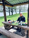 Älterer Mann sitzt an überdachtem Holztisch mit Picknick am Seeufer in grüner Landschaft.