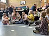 Kinder sitzen auf dem Hallenboden vor einer Tafelrunde mit Erwachsenen in einer Turnhalle.