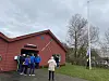 Gruppe von Menschen vor einem roten Bootshaus mit Flaggenmast auf einer Wiese