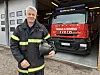 Älterer Feuerwehrmann in Einsatzjacke mit Helm steht vor rotem Iveco-Feuerwehrfahrzeug im Gerätehaus.