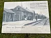 Infotafel mit historischem Foto eines Bahnhofs neben Gleisen auf einer Rasenfläche.