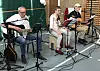 Drei Musiker mit Gitarren und Mikrofonen spielen in einer Turnhalle auf einer kleinen Bühne.