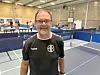 Älterer Mann in Sportkleidung steht lächelnd in einer Tischtennishalle vor mehreren Tischen.