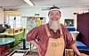 Künstler mit Bart in Latzhose steht lächelnd in einem bunten Atelier vor seinen Arbeiten.