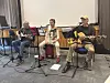 Drei Musiker mit Gitarren und Mikrofonen musizieren in einer Aula auf kleiner Bühne.