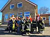 Kinder in Feuerwehrschutzkleidung stehen mit Erwachsenen vor einem Schulgebäude im Freien.
