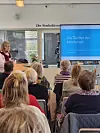 Publikum in einer Bibliothek bei einem Vortrag vor Leinwand mit dem Titel „Die Töchter der Minderheit“.
